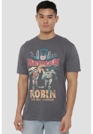 Junger Mann trägt ein graues Batman- und Robin-Grafik-T-Shirt mit laufenden Figuren und Stadtsilhouette im Hintergrund.
