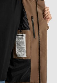 Hnedá bunda s hladkým vonkajším materiálom, čiernou podšívkou a zipsovým vreckom. Čitelný štítok vo vnútri obsahuje pokyny na údržbu a značku.