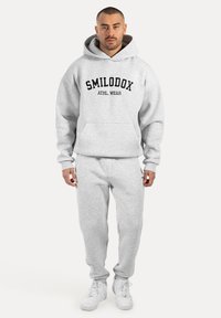Ensemble en hoodie gris et pantalon de survêtement, avec poche avant et logo en texte noir sur le hoodie. Tissu doux, coupe décontractée, associé à des baskets blanches.
