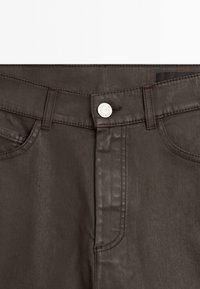 Brown denim pants featuring a silver button closure, visible stitching, and side pockets. The fabric has a smooth, slightly shiny texture.