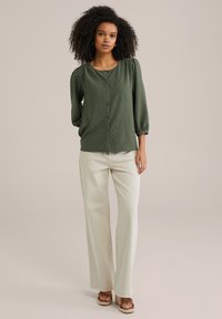 Blusa a maniche lunghe verde a pois con scollatura tonda, caratterizzata da bottoni sulla parte anteriore. Abbinata a pantaloni a gamba larga di colore chiaro.