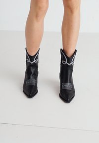 Black faux leather cowboy boots with pointed toes, embroidered accents, and folded cuffs; features white stitching and textured pattern.