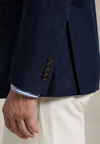 Tengerészkék vászon blézer egy hátsó sliccel, négy sötét gombbal az ujján, alatta világoskék-fehér csíkos ing.