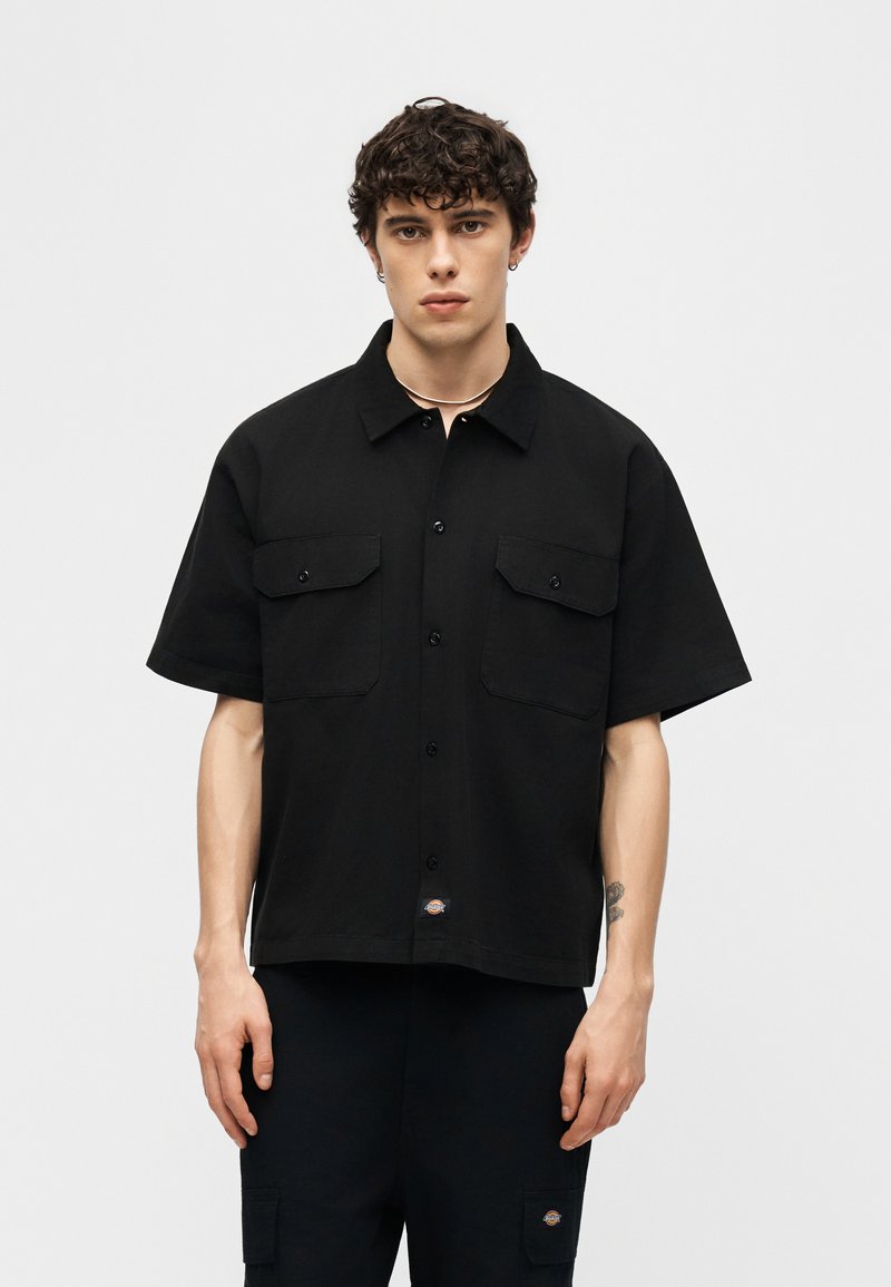 Giovane uomo con capelli ricci che indossa una camicia nera a maniche corte con bottoni e tasche sul petto, abbinata a pantaloni neri.