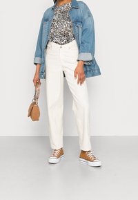 Personne portant une veste en denim bleu, un haut imprimé animal, un pantalon blanc taille haute, des baskets montantes marron et tenant un sac à main beige.