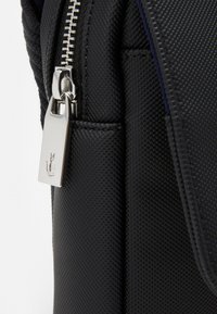 Svart teksturert stoffbag med en synlig metallglidelås og drasnor som har en liten logo. Nærbilde av sømmen og skjøtene.