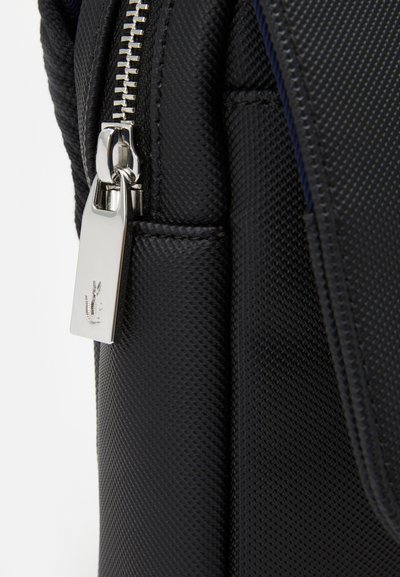 Sac en tissu noir texturé avec une fermeture éclair métallique et un tirette visibles, arborant un petit logo. Gros plan sur les coutures et les joints.