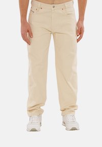 Beige Baumwollhose mit geradem Schnitt, ausgestattet mit frontalen Taschen, metallischen Hardware-Elementen und einer subtilen Textur. Kombiniert mit weißen Sneakers.