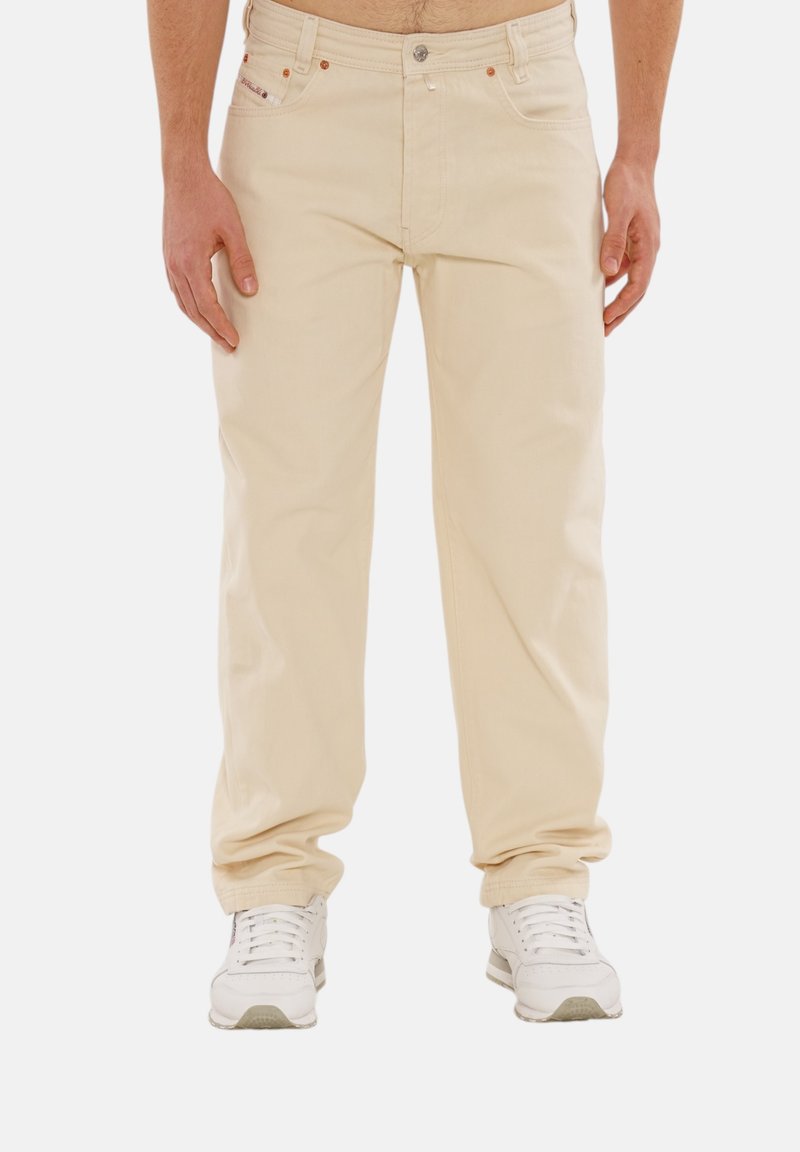 Beige Baumwollhose mit geradem Schnitt, ausgestattet mit frontalen Taschen, metallischen Hardware-Elementen und einer subtilen Textur. Kombiniert mit weißen Sneakers.
