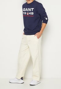 Marineblauwe sweatshirt met witte en rode tekst, crèmekleurige broek, en witte sneakers met marineblauwe accenten; casual pasvorm en zachte stof.