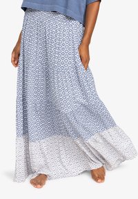 Mujer vistiendo una falda larga de patrones azules y blancos con bolsillos, descalza sobre un fondo blanco.