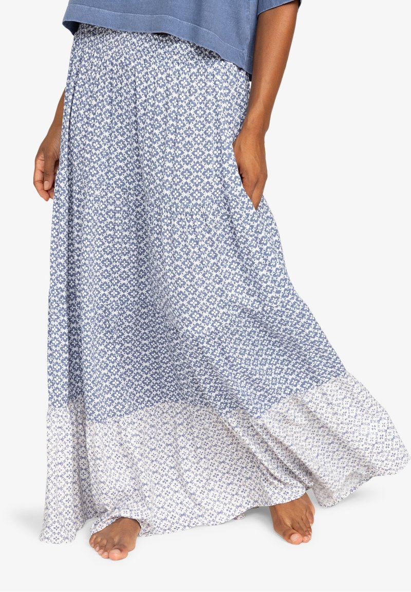 Mujer vistiendo una falda larga de patrones azules y blancos con bolsillos, descalza sobre un fondo blanco.