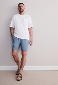 Maglietta bianca a maniche corte con tasca, pantaloncini azzurri e sandali marroni con cinturini regolabili, in piedi su uno sfondo neutro.