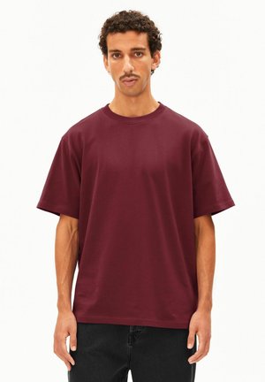 Mann mit lockigem Haar und Schnurrbart, der ein einfarbiges dunkelrotes Kurzarmhemd und schwarze Hosen vor weißem Hintergrund trägt.