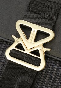 Zwarte tasriem met een grote gouden metalen gesp met een geometrisch ontwerp, tegen een achtergrond van zwart textiel.