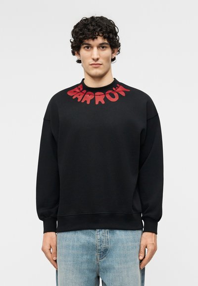 Νεαρός άνδρας με σγουρά μαλλιά φοράει μαύρο φούτερ με κόκκινο κείμενο "BARROW" στον γιακά, συνδυασμένο με ανοιχτόχρωμα μπλε τζιν, στέκεται μπροστά σε απλό φόντο.