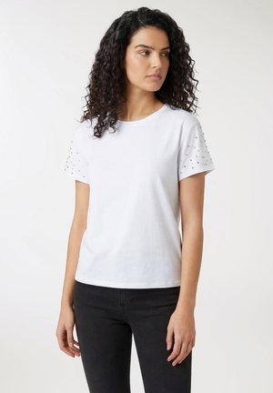 Donna con capelli ricci e scuri che indossa una maglietta bianca a maniche corte con borchie argento sulle maniche e pantaloni neri, che guarda verso sinistra.