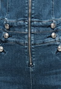 Τζιν ολόσωμη φόρμα σε σκούρο μπλε με εφαρμοστό σχέδιο. Διαθέτει ασημί κουμπιά και φερμουάρ μ προστά. Υφή με φθορές κατά μήκος των ραφών.