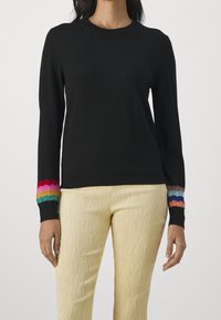 Kobieta ubrana w czarny sweter z długim rękawem, z kolorowymi paskami u mankietów i teksturowanymi, jasnobeżowymi spodniami, stoi na tle jednolitym.