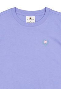 Lila krótki rękaw T-shirt wykonany z bawełny, z małym emblematem w kształcie kwiatu z liśćmi po lewej stronie klatki piersiowej. Okrągły dekolt i miękka faktura.