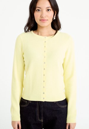 Kvinde iført en lysegul tekstureret cardigan med knapper og mørkeblå jeans, stående foran en ensfarvet hvid baggrund.