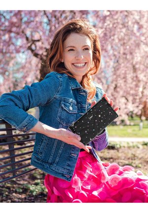 Black wallet with multi-colored polka dots, zip closure, and quilted texture, held by a person in a denim jacket against a floral background.