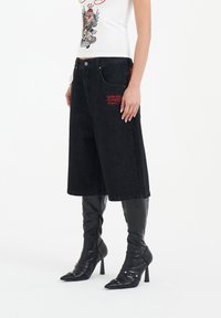 Musta farkkuculottes, joissa on leveälahkeinen muotoilu, kirjailtua brändäystä taskussa, yhdistettynä mustiin polven yli ulottuviin stiletto-korkokenkiin.
