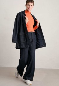 Giacca di jeans blu scuro con grandi tasche, abbinata a un dolcevita arancione aderente e pantaloni di jeans a gamba larga. Le sneakers completano l'outfit.
