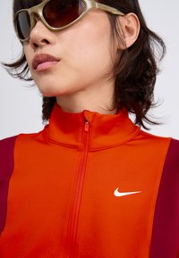 Genser med glidelås i oransje og mørkerød, med høy krage og en hvit Nike-logo. Glatt stoff med lett tekstur, kombinert med solbriller.