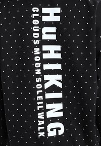 Svart tyg med små vita trianglar och fet vit vertikal text som säger "HUIHKING" och mindre text "CLOUDS MOON SOLEIL WALK."