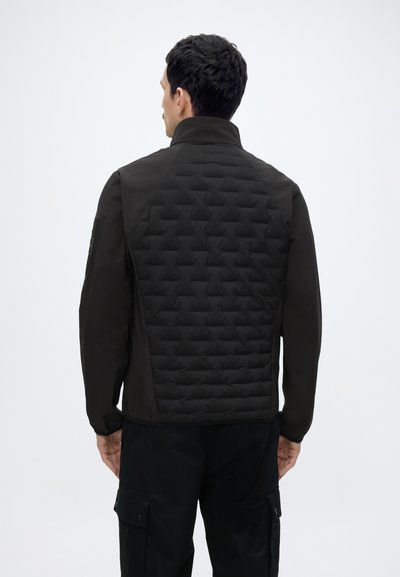 Homme de dos portant une veste matelassée noire à col relevé et un pantalon cargo noir sur fond blanc uni.