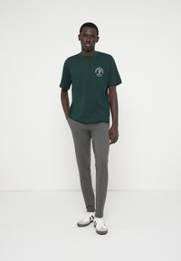 Tmavě zelené bavlněné tričko s kulatým výstřihem a krátkými rukávy, s bílým grafickým potiskem. V kombinaci s úzkými šedými kalhotami a teniskami.