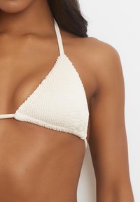 Vit bikiniöverdel med strukturerade triangelkoppar och tunna band. Materialet har ett vågigt mönster längs ytan.