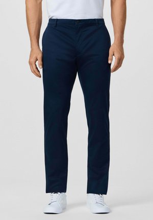 Man wearing straight-fit navy blue trousers and white sneakers, standing against a plain light background.