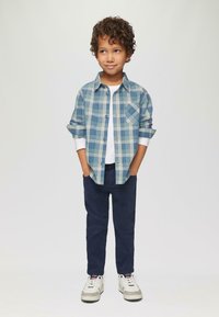 Geruite overhemd in blauwe en groene tinten, witte T-shirt, marineblauwe broek en witte sneakers. Het overhemd heeft opgerolde mouwen en een pocket.