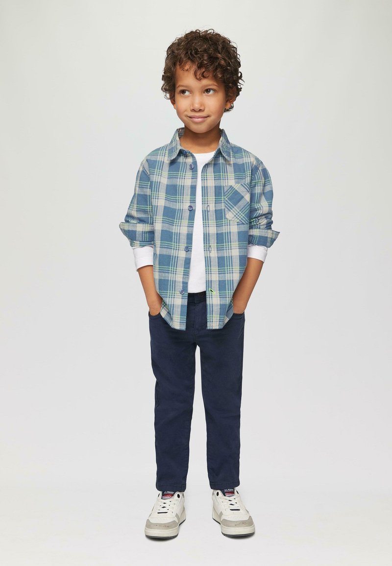 Geruite overhemd in blauwe en groene tinten, witte T-shirt, marineblauwe broek en witte sneakers. Het overhemd heeft opgerolde mouwen en een pocket.