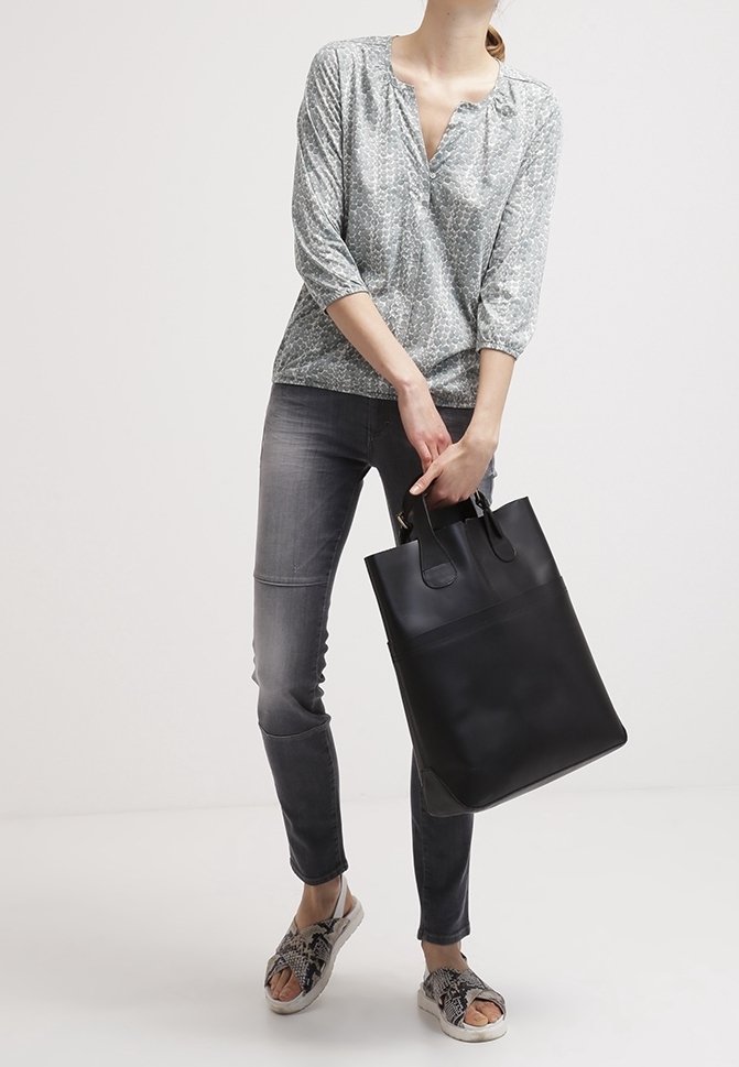 Femme portant un haut gris à motifs et un jean foncé, tenant un grand sac fourre-tout noir, debout les jambes croisées et portant des sandales en cuir de serpent.