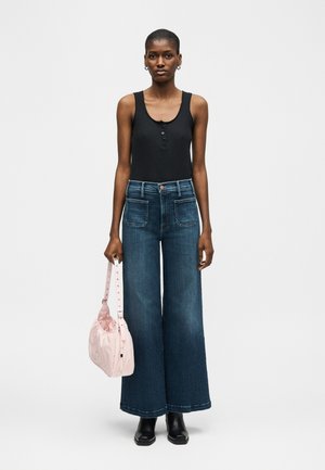 Γυναίκα που φοράει μαύρο αμάνικο τοπ και φαρδιά μπλε τζιν, κρατώντας ένα ανοιχτό ροζ τσαντάκι στο αριστερό της χέρι, στέκεται σε απλό φόντο.