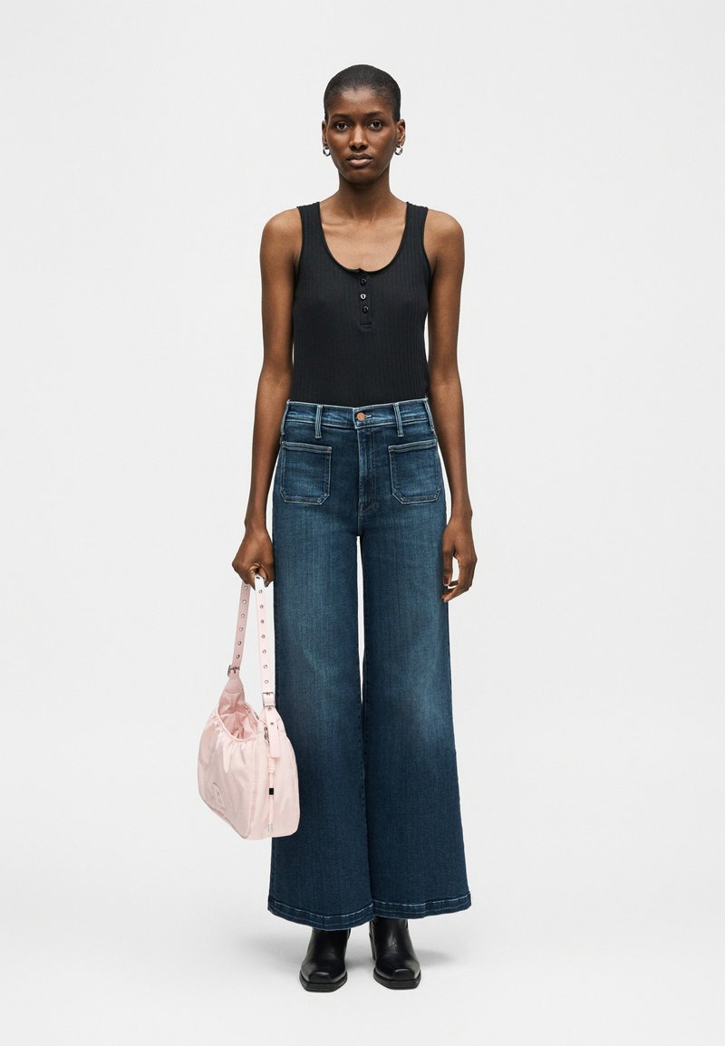 Γυναίκα που φοράει μαύρο αμάνικο τοπ και φαρδιά μπλε τζιν, κρατώντας ένα ανοιχτό ροζ τσαντάκι στο αριστερό της χέρι, στέκεται σε απλό φόντο.