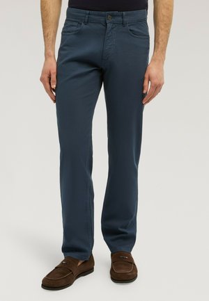 Mann trägt marineblaue, gerade geschnittene Hose und braune Wildleder-Loafers und steht vor einem schlichten Hintergrund.