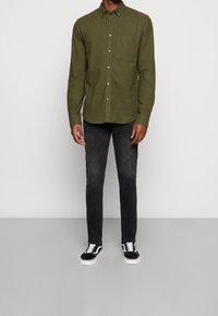 Uomo in camicia verde oliva a bottoni, jeans neri slim-fit e sneakers bianco e nero che si trova davanti a uno sfondo neutro.