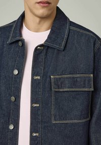 Veste en denim bleu foncé, avec des coutures claires, des boutons en métal et une poche en patch sur le côté gauche, portée par-dessus une chemise rose clair.