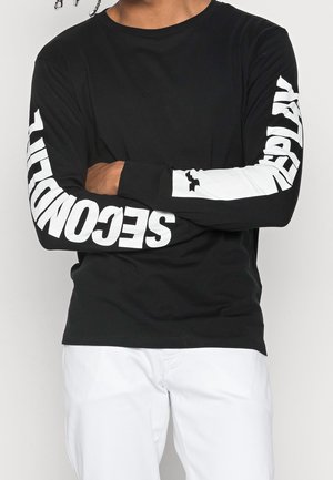 Personne portant un haut noir à manches longues avec le texte blanc « SECOND » sur la manche gauche et « REPLAY » sur la manche droite, associé à un pantalon blanc.