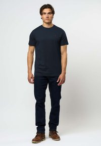 Mladić stoji, nosi jednobojnu tamnoplavom majicu kratkih rukava, tamne traperice i smeđe kožne čizme, na jednostavnoj svijetloj pozadini.