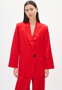 Roter Blazer aus glattem Stoff, ausgestattet mit einem einzelnen schwarzen Knopf, Reverskragen und weitem Ärmel. Kombiniert mit passender Hose.