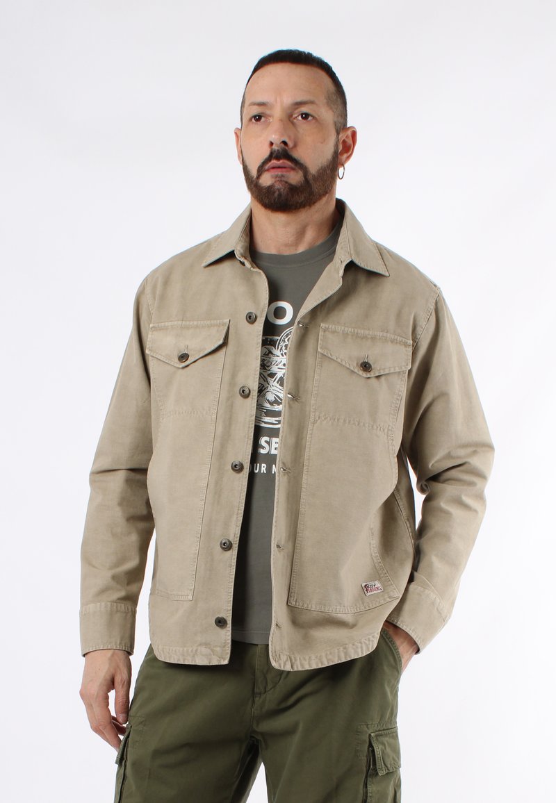 Giacca in cotone beige con chiusura a bottoni, due tasche sul petto e una vestibilità morbida. Abbinata a una maglietta grigia con graphic e pantaloni cargo verdi.