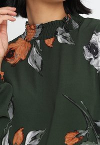 Vihreä pusero, jossa on oranssia ja harmaata kukkakuosi. Ominaisuuksiin kuuluu rypytetty kaula-aukko ja pitkät puffihihat. Silkkinen, kevyt materiaali.