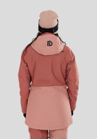 Roze waterdichte jas met een capuchon, bicolor ontwerp en logo op de achterkant. Het model draagt een lichtroze muts en zwarte handschoenen.