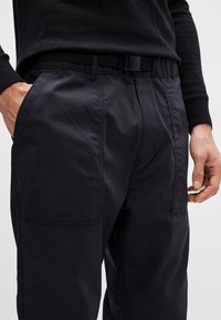 Calças azul marinho escuras, feitas de um tecido leve, com um corte descontraído, bolsos laterais e um cinto preto ajustável. Textura lisa com um brilho subtil.