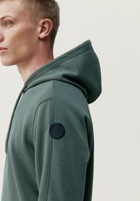 Mikina s kapucňou tmavozelenej farby, s okrúhlym čiernym logom na ľavom pleci; hladká textúra a priliehavý strih.